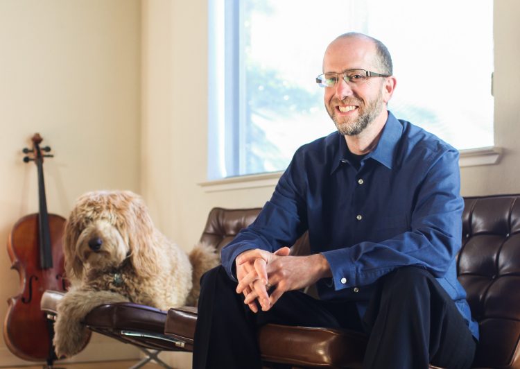 Matt Eisenberg sitting on a brown leather couch next to his Bearded Collie.